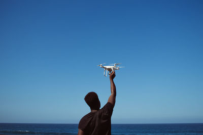 Rear view of man holding drone against blue sky