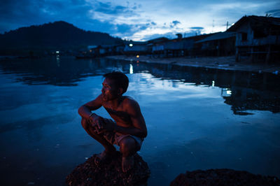 Young man sitting on rock by lake against sky