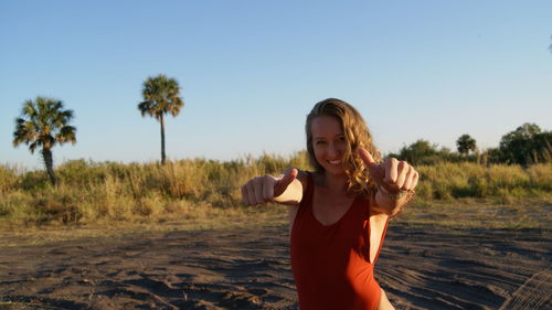 Portrait of cheerful young woman showing thumbs up at beach against clear sky