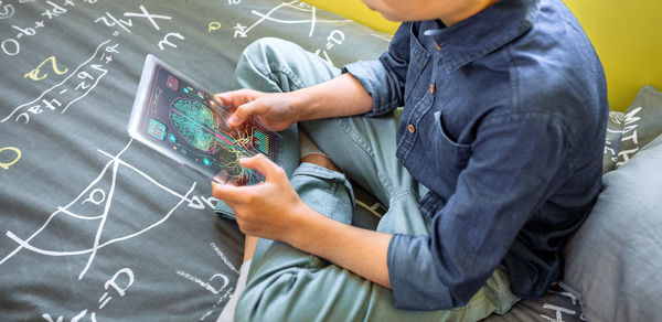 Unrecognizable young kid holding digital display with glass screen to study sitting over his bed