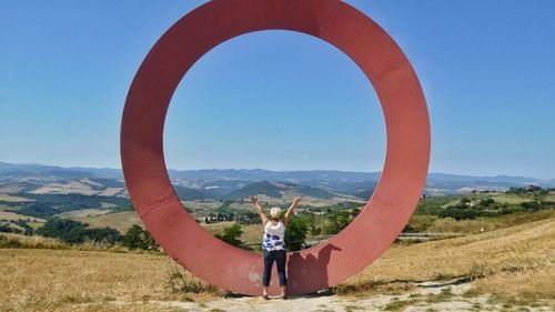 Full length rear view of woman with arms raised standing on landscape by circle shaped built structure against clear sky