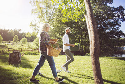 Full length of friends holding vegetable crate and basket while walking on grassy field