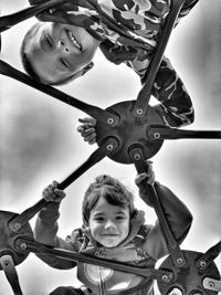 Low angle view portrait of happy siblings on monkey bars against cloudy sky