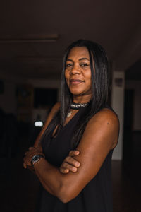 Attractive african american female with long dark hair in black outfit standing with crossed arms in dark room and looking at camera