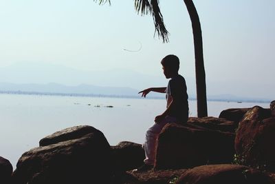 Boy sitting on rock by sea against sky