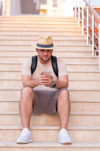 Handsome adult blond bearded man wearing sunhat, backpack using cellphone, mobile, texting, sitting