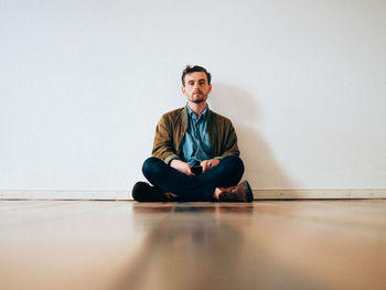 Full length portrait of serious man sitting on hardwood floor against white wall