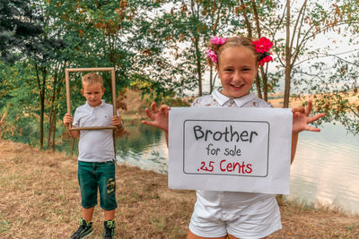 Portrait of smiling girl holding information sign while standing with brother against lake