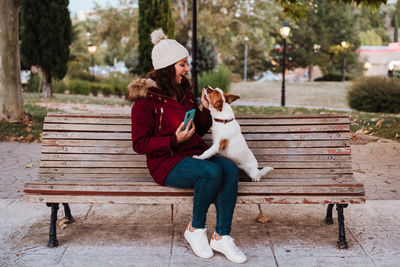 Woman with dog using phone while sitting on bench against trees