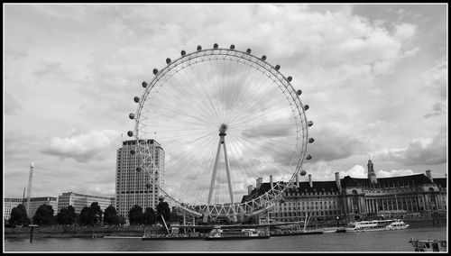 Ferris wheel on riverbank against cloudy sky