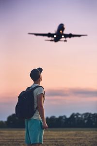 Side view of man with backpack looking at airplane against sky during sunset