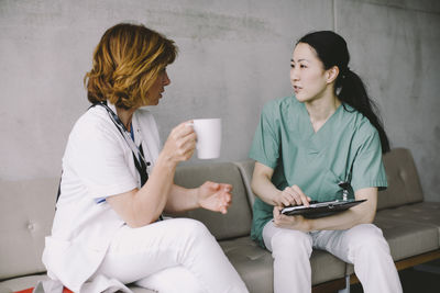 Senior doctor holding coffee cup while discussing with nurse sitting on sofa at hospital