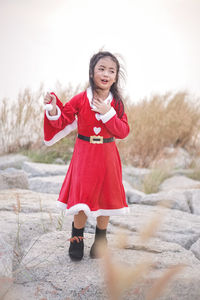 Cute girl wearing red dress holding santa hat while standing on rocks against sky