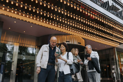 Male and female senior friends discussing with each other while using smart phones outside movie theater