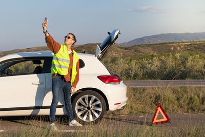 Young man on vest close to his broken white car trying to get and find signal on the phone 