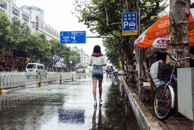 Full length rear view of woman walking on wet city street against sky