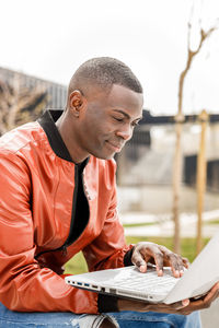 Side view of concentrated happy young african american male freelancer in trendy outfit sitting on stone bench and working remotely on laptop in city park