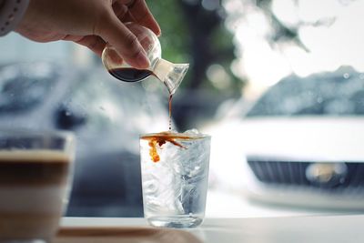 Cropped hand of woman pouring coffee in cold drink by window at cafe