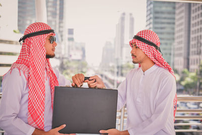 Man giving briefcase to friend standing at bridge in city