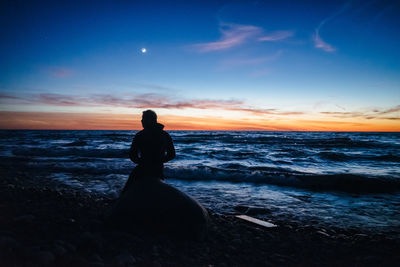 Man sitting on beach against sky during sunset