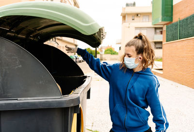 Girl throwing the a yellow trash bag into a green recycling container open in the street. the teenager is wearing face mask and gloves to protect herself from infections, virus, bacterias. ho