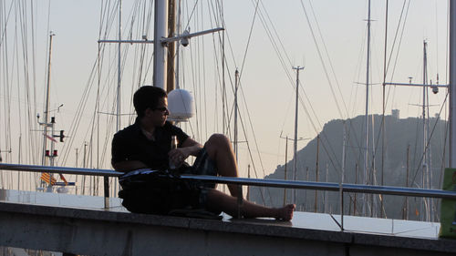 Full length of man with beer bottle resting on retaining wall by moored sailboats during sunset