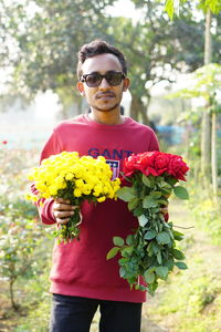 Portrait of a young man wearing sunglasses standing holding flowers in both hands