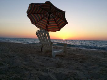 Empty chair by beach umbrella at shore of beach during sunset 