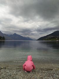 Rear view of child crouching at lakeshore against cloudy sky