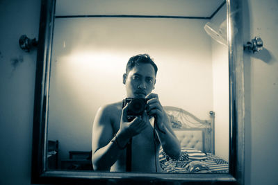 Reflection of shirtless man photographing through camera in mirror at home