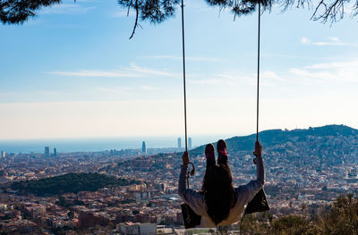 Rear view of woman swinging on rope swing against cityscape and sky