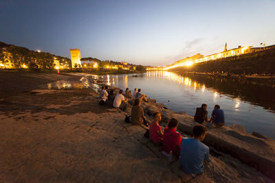 People on riverbank against sky at sunset