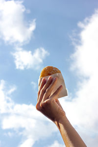 Low angle view of hand holding donut bread against sky