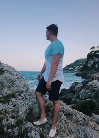 Full length of young man standing on rock by sea against clear sky