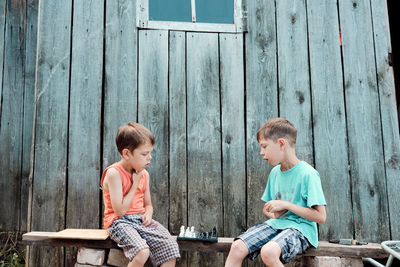 Two village boys play chess on the street, sitting on a bench, against the backdrop of an old barn