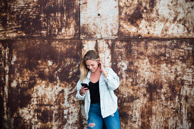 Young woman using phone while standing against wall