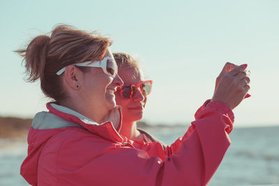 Side view of mother photographing sea while standing with daughter against sky during sunset