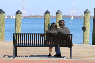Rear view of couple sitting at riverbank against clear sky