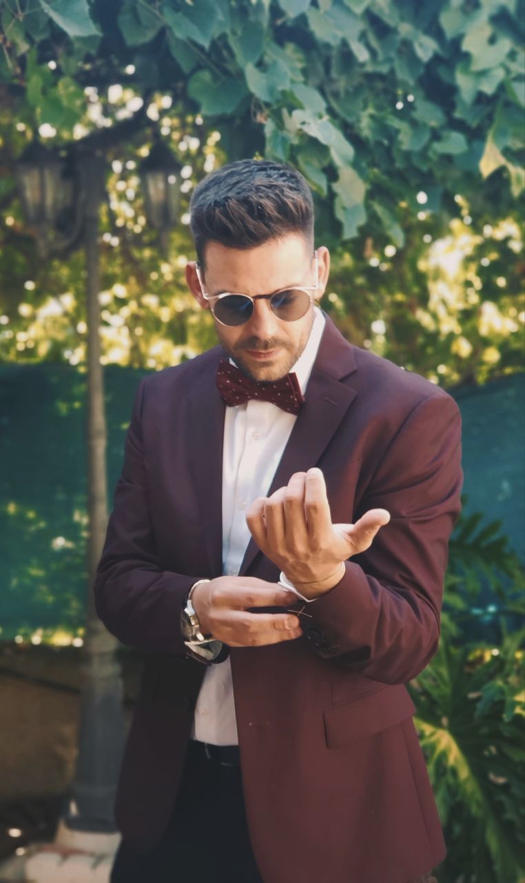 one person, young adult, front view, standing, young men, real people, fashion, three quarter length, sunglasses, focus on foreground, well-dressed, day, glasses, business, formalwear, sunlight, outdoors, suit