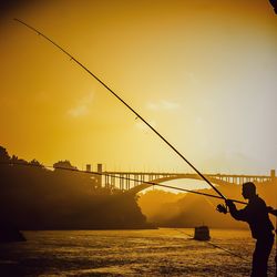 Silhouette man fishing on bridge against sky during sunset