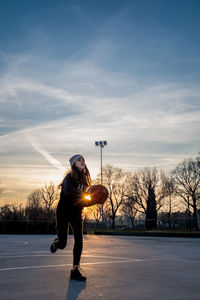 Young girl practices alone in the game of basketball. outdoor playground with low setting sun.