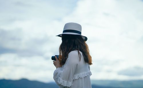 Woman holding camera while standing against cloudy sky
