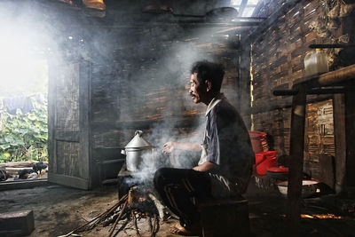 Side view of man sitting by wood burning stove in hut