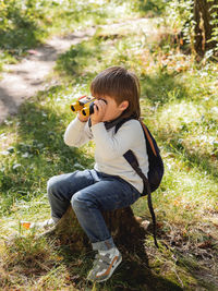 Little explorer on hike in forest. boy with binoculars sits on stump. outdoor leisure for children. 