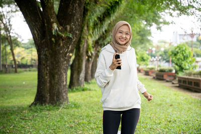 Full length of smiling young woman using phone while standing on tree
