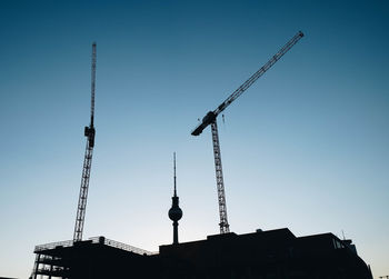 Low angle view of silhouette crane by building against clear blue sky