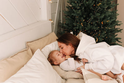 Smiling sleepy mom and baby wake up lying on the bed by the christmas tree in the bedroom at home