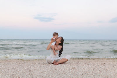 Mother kissing daughter while sitting on shore at beach against sky