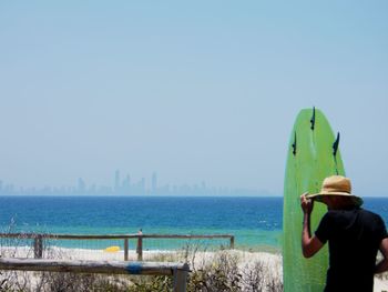 Rear view of man wearing hat standing with surfboard against sea and sky