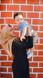 Mother lifting son while standing against brick wall